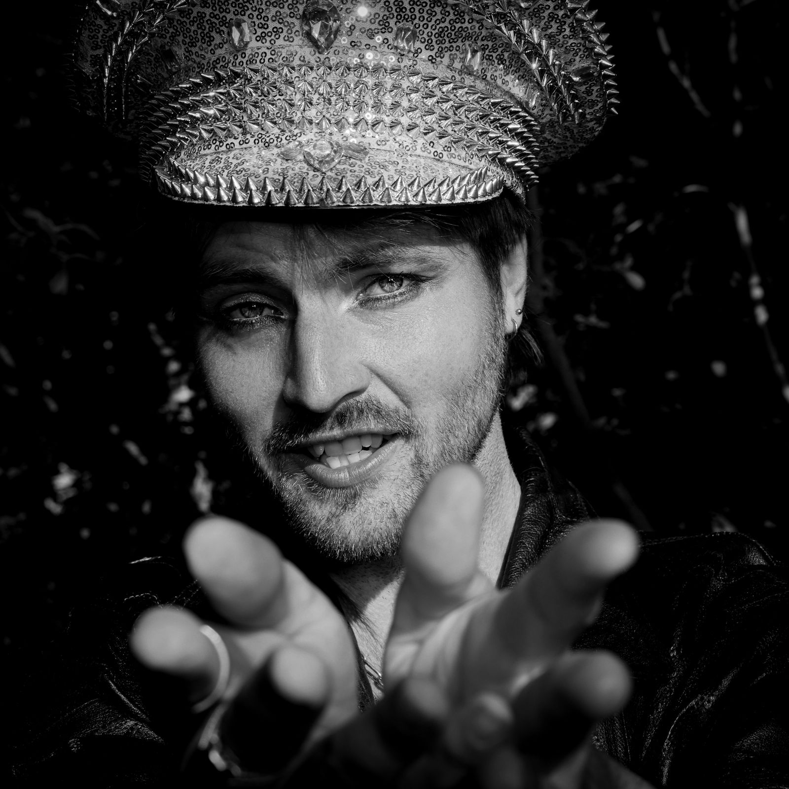 Johan looks directly into the camera, wearing an ornate cap and dark clothing, with his hand reaching toward the viewer in a playful, intimate portrait.