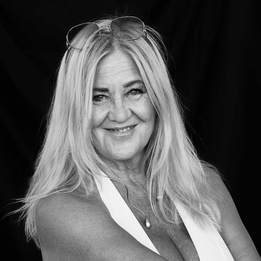 Portrait of a smiling woman with long blonde hair, wearing sunglasses on her head, against a black background.