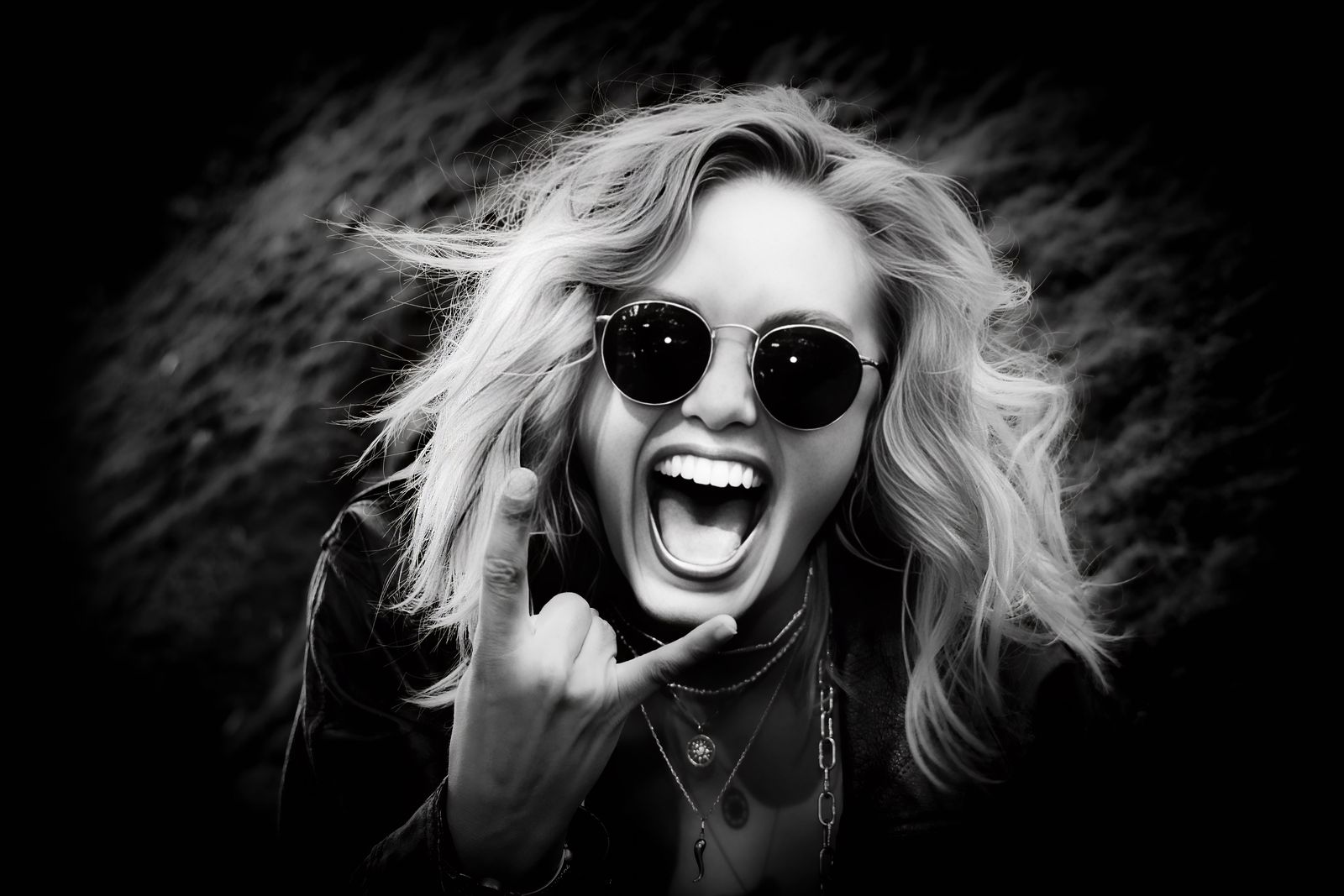 Black-and-white portrait of Mira laughing with her mouth wide open, wearing round dark sunglasses and layered necklaces, playfully making a shaka hand gesture against a dark natural background.